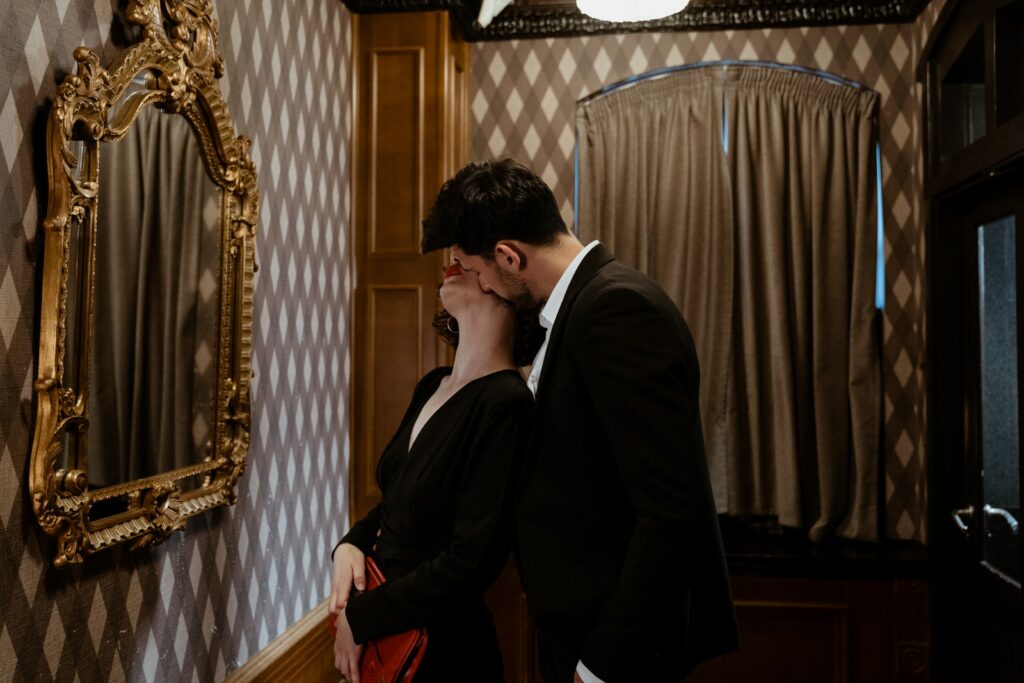 Man in Black Suit Kissing Woman in Black Dress Kissing on the Neck in Front of Mirror